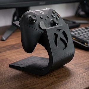 Könnte beinhalten: Ein schwarzer Xbox-Controller liegt auf einem passenden schwarzen Ständer. Der Ständer hat ein geschwungenes Design mit ausgeschnittenem Xbox-Logo. Der Controller befindet sich auf einem Holzschreibtisch in der Nähe eines Computermonitors und einer Tastatur.