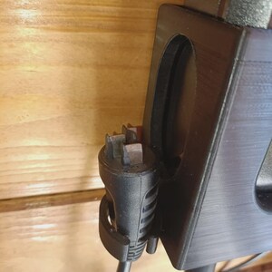 Puede incluir: Un soporte de pared de pl&aacute;stico negro con una ranura vertical y un clip circular. Un enchufe el&eacute;ctrico negro con cuatro clavijas met&aacute;licas est&aacute; asegurado en el clip, mientras que un cable de alimentaci&oacute;n negro cuelga del enchufe.