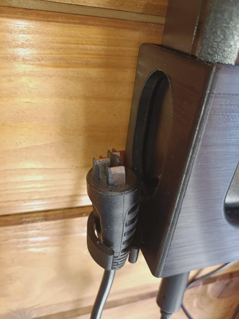 Puede incluir: Un soporte de pared de pl&aacute;stico negro con una ranura vertical y un clip circular. Un enchufe el&eacute;ctrico negro con cuatro clavijas met&aacute;licas est&aacute; asegurado en el clip, mientras que un cable de alimentaci&oacute;n negro cuelga del enchufe.