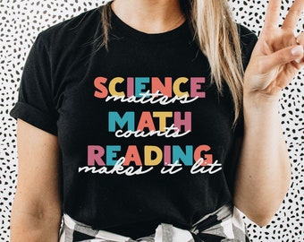May include: A black short-sleeved t-shirt featuring colorful text that reads SCIENCE matters, MATH counts, and READING makes it lit. The words are printed in a mix of pink, yellow, and blue block letters and white cursive script on the front.