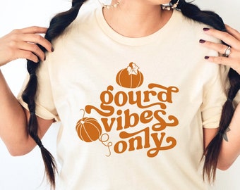 May include: Cream-colored t-shirt with the phrase "gourd vibes only" in orange lettering. The design includes two pumpkin illustrations. A fall-themed graphic tee for autumn and Halloween.
