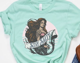 May include: A turquoise t-shirt with a graphic of a mermaid with long brown hair and a tail. The mermaid is holding a banner that says "Stay Salty".