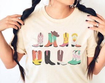 May include: A beige t-shirt with a graphic of colorful cowboy boots. The boots are arranged in a row and have different designs, including a heart, a star, and a cowboy hat.