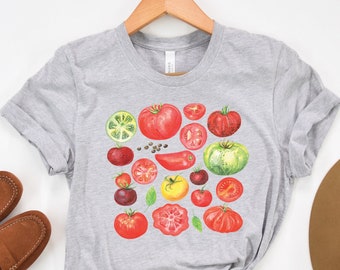 Puede incluir: Una camiseta gris con una ilustración colorida de diferentes rodajas de tomate, tomates enteros y pimientos verdes. La ilustración está centrada en la camiseta.