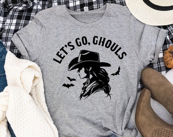 May include: A gray t-shirt with the text "Let's Go, Ghouls" in black. A black and white illustration of a woman wearing a cowboy hat and a long-sleeved shirt is below the text. The t-shirt is laid out on a white surface with a plaid shirt, a white hat, a pair of jeans, a white sweater, and a pair of brown boots.