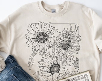 Puede incluir: Una sudadera beige con un dibujo en blanco y negro de girasoles en la parte delantera. La sudadera está sobre una superficie blanca con un par de zapatillas blancas, unos vaqueros azules y un libro.