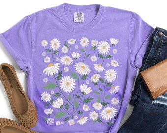 May include: A purple t-shirt with a white daisy floral print. The shirt is paired with blue jeans and brown shoes.