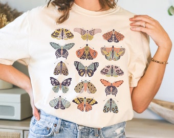 May include: Cream-colored t-shirt featuring a collection of colorful butterfly illustrations. The butterflies are in various shapes and sizes, with detailed wing patterns in shades of yellow, blue, orange, and purple. The shirt is made of a soft material.