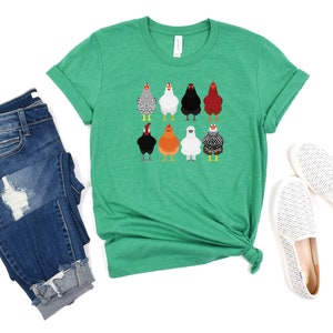 Puede incluir: Una camiseta verde con un gr&aacute;fico de ocho pollos diferentes. Los pollos son de diferentes colores y razas. La camiseta est&aacute; doblada y colocada sobre un fondo blanco.