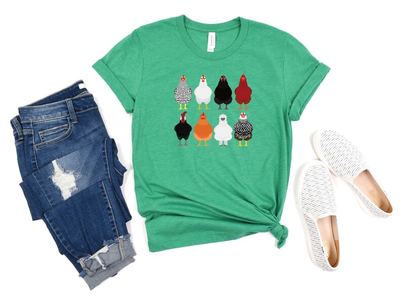Puede incluir: Una camiseta verde con un gr&aacute;fico de ocho pollos diferentes. Los pollos son de diferentes colores y razas. La camiseta est&aacute; doblada y colocada sobre un fondo blanco.