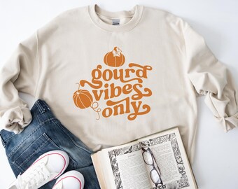 May include: A beige crewneck sweatshirt featuring orange pumpkin graphics and the text "gourd vibes only" in a cursive font. The sweatshirt is paired with blue denim jeans, white sneakers, and an open book with black-rimmed reading glasses.