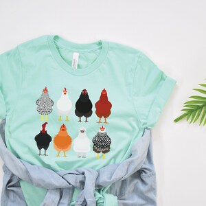 Puede incluir: Una camiseta azul claro con un gr&aacute;fico de ocho pollos diferentes en varios colores y patrones. Los pollos est&aacute;n todos mirando hacia adelante y tienen la cabeza levantada.
