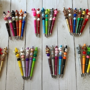 Peut inclure: Une collection de stylos à bille avec des embouts en silicone représentant des personnages et des perles décoratives. Chaque stylo possède une figurine unique d'animal ou de créature, incluant des ours, renards, abeilles et pandas, avec des corps de différentes couleurs.