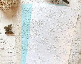Könnte beinhalten: Geprägtes Papier mit Schneeflockenmustern in Weiß, Hellblau und Creme. Das Papier hat eine strukturierte Oberfläche mit erhabenen Schneeflockenmustern. Dekorative Metallakzente in Form von Schmetterlingen und Libellen sind sichtbar.