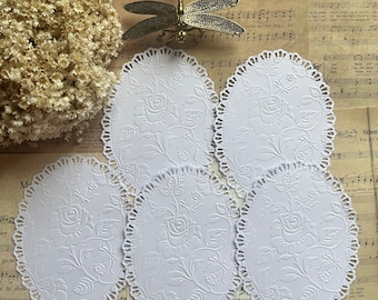 Puede incluir: Cinco servilletas de papel ovaladas blancas con un borde de encaje delicado y un diseño floral en relieve. Las servilletas están dispuestas en círculo sobre un fondo de partituras musicales antiguas.