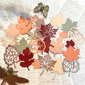 May include: An assortment of paper leaves in autumn shades of orange, red, and brown. Some leaves feature detailed cut-out patterns. A decorative brass butterfly and dragonfly are also visible.