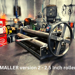 May include: A metal roller for bending sheet metal. The roller has a 2.5 inch diameter and is made of steel. The roller is mounted on a metal frame with a hand crank. The roller is in a workshop setting.