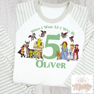 May include: White and grey striped long-sleeved shirt with a green graphic featuring Shrek characters, the number 5, and the text "When I Wake Up I Will Be 5 Oliver."