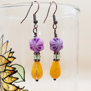Puede incluir: Pendientes colgantes de color morado y amarillo con una cuenta en forma de flor y una cuenta en forma de lágrima.