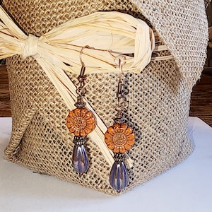 May include: A pair of dangle earrings with a copper wire and brown and orange ceramic flower accents. The flower accents have a textured surface and the bottom of the earrings are a dark blue color.