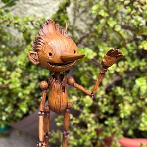 Puede incluir: Una figurita de madera de un personaje fantasioso con una nariz larga, ojos grandes y una sonrisa amigable. La figurita está articulada y tiene una textura de veta de madera.