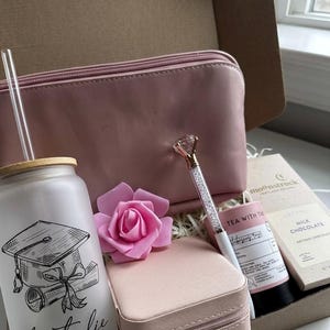 May include: A pink-themed gift box with a graduation glass, a cosmetic bag, a pen with a diamond-shaped top, a small jewelry case, a pink rose, tea, and chocolate. The glass is personalized.
