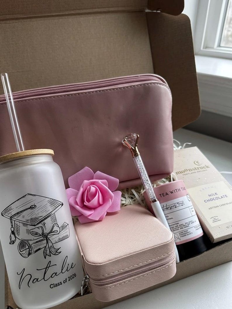 May include: A pink-themed gift box with a graduation glass, a cosmetic bag, a pen with a diamond-shaped top, a small jewelry case, a pink rose, tea, and chocolate. The glass is personalized.