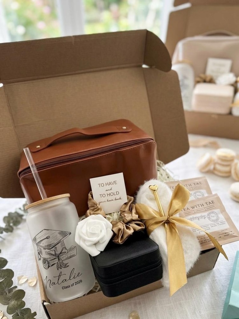 May include: A gift box containing a brown leather-look travel case, a frosted glass tumbler with a straw featuring a graduation cap graphic and the text Natalie Class of 2026, a black jewellery box, a white rose, gold scrunchies, and tea.