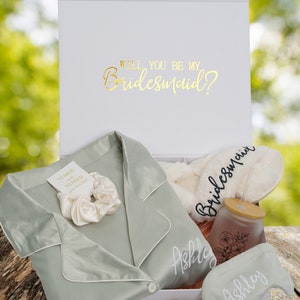 May include: A white gift box with a green satin robe, a white satin scrunchie, a white fluffy slipper with the word "Bridesmaid" embroidered on it, a glass jar with a bamboo lid and a straw, and a sleep mask with the word "Ashley" embroidered on it. The box lid has the text "Will you be my Bridesmaid?" in gold lettering.