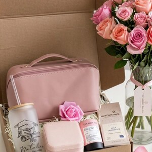 May include: A gift box containing a pink wash bag, a tumbler with a graduation cap design, a jewellery box, a rose, a tube of tea, and a bar of milk chocolate. A bouquet of pink and peach roses is in a glass vase.