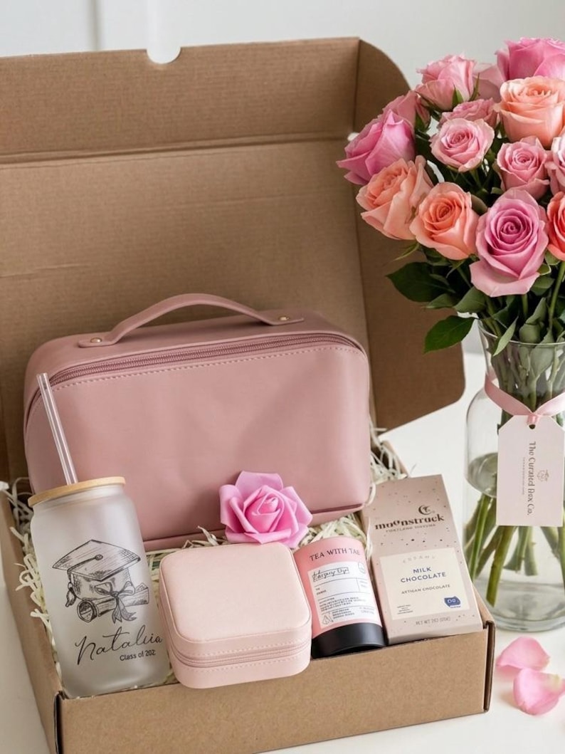 May include: A gift box containing a pink wash bag, a tumbler with a graduation cap design, a jewellery box, a rose, a tube of tea, and a bar of milk chocolate. A bouquet of pink and peach roses is in a glass vase.