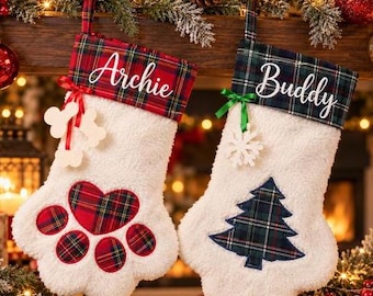 May include: Two white plush paw-shaped Christmas stockings with plaid cuffs. The left stocking features a red plaid heart-shaped paw pad, a red ribbon with bone-shaped tags, and the name Archie. The right stocking has a green plaid tree, a green ribbon with a snowflake, and the name Buddy.