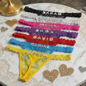 May include: A collection of colorful thong underwear with lace trim and rhinestone lettering spelling out names and zodiac signs. The underwear is in various colors, including black, white, pink, purple, red, blue, and yellow. Decorative heart-shaped embellishments are scattered around.
