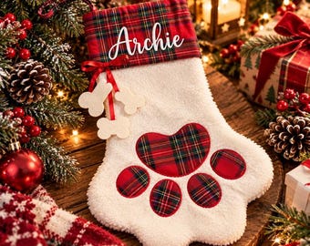 May include: A white plush paw-shaped Christmas stocking featuring a red and green plaid cuff with the name Archie in white script. The stocking includes a red ribbon with three small bone-shaped ornaments and a plaid paw pad design.