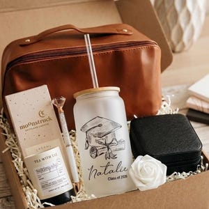May include: A gift box containing a brown leather-like pouch, a frosted glass tumbler with a bamboo lid, a clear straw, and a graduation graphic with the text Natalie Class of 2026. Includes a white pen with a diamond topper, a candle, and a black case.