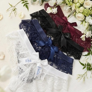 May include: Four lace garters in white, navy, black, and burgundy, each with a satin ribbon bow. The white garter has "Mrs. Porter" printed on a ribbon. A floral arrangement is in the background.