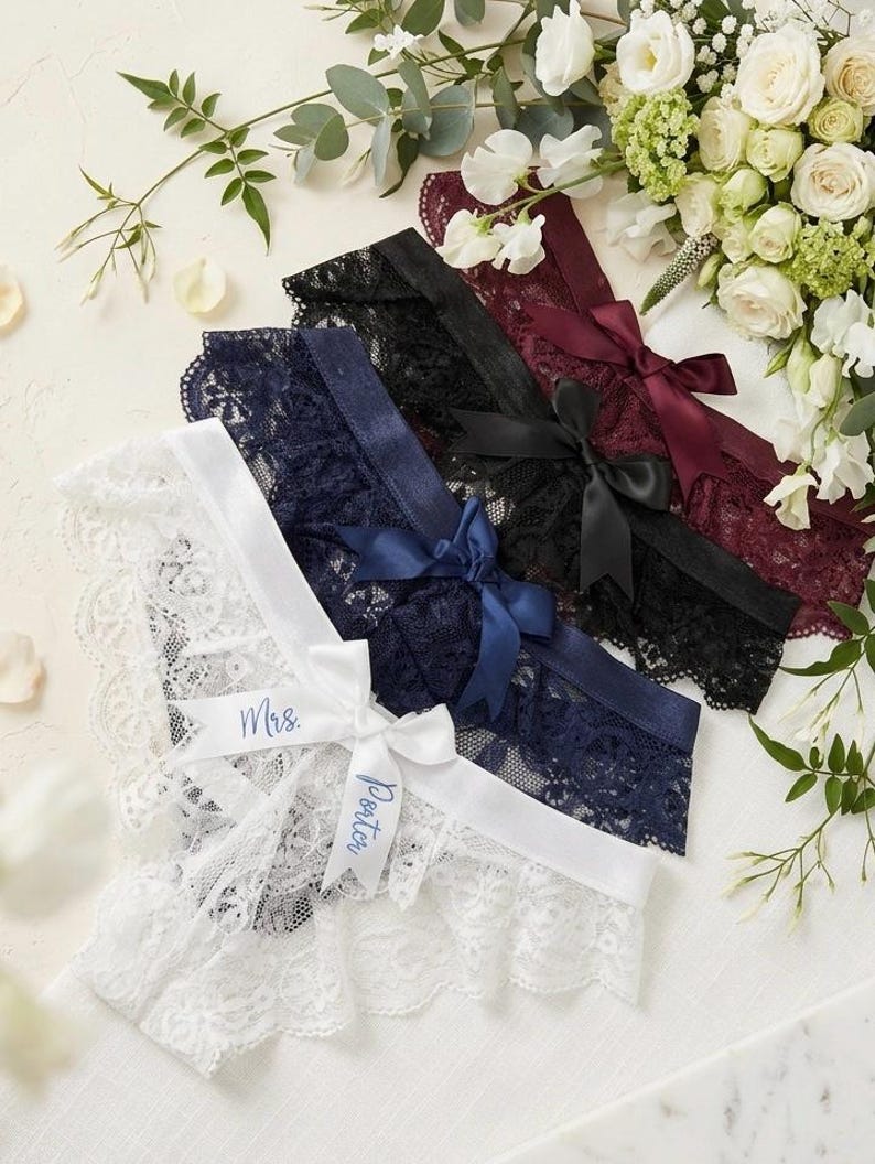 May include: Four lace garters in white, navy, black, and burgundy, each with a satin ribbon bow. The white garter has "Mrs. Porter" printed on a ribbon. A floral arrangement is in the background.