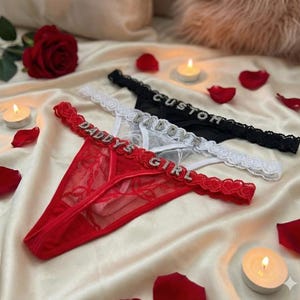 Peut inclure: Trois strings en rouge, blanc et noir. Le string rouge porte l'inscription "DADDY'S GIRL" en strass. Le string blanc porte l'inscription "DADDY" en strass. Le string noir porte l'inscription "CUSTOM" en strass. Bordure en dentelle.