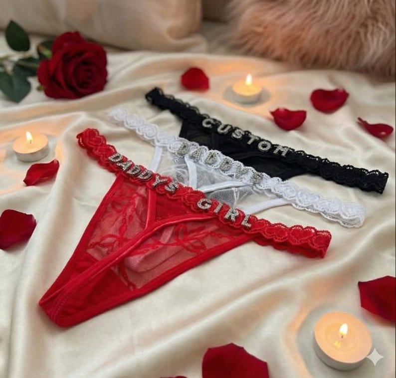 Peut inclure: Trois strings en rouge, blanc et noir. Le string rouge porte l'inscription "DADDY'S GIRL" en strass. Le string blanc porte l'inscription "DADDY" en strass. Le string noir porte l'inscription "CUSTOM" en strass. Bordure en dentelle.