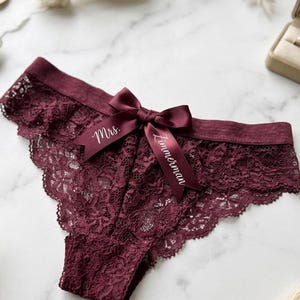 May include: Burgundy lace underwear with a satin bow. The bow is inscribed with the words "Mrs. Zimmerman" in white script. The underwear has a scalloped edge and is displayed on a marble surface.