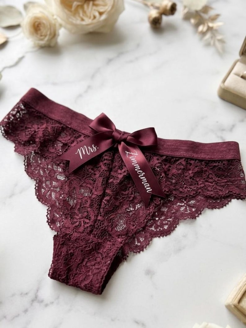May include: Burgundy lace underwear with a satin bow. The bow is inscribed with the words "Mrs. Zimmerman" in white script. The underwear has a scalloped edge and is displayed on a marble surface.