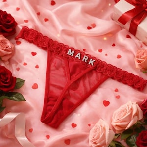 Peut inclure: String rouge avec le nom "MARK" en strass. Le sous-v&ecirc;tement est orn&eacute; d'une d&eacute;licate bordure en dentelle et est pr&eacute;sent&eacute; sur une surface en satin rose avec des confettis en forme de c&oelig;ur et des roses.