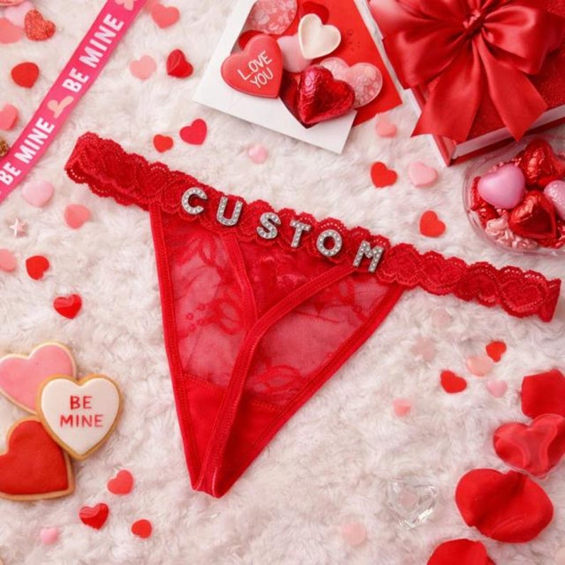 Peut inclure: String en dentelle rouge avec le mot "CUSTOM" en lettres argent&eacute;es. L'image comprend des bonbons, des biscuits et une bo&icirc;te cadeau en forme de c&oelig;ur, tous rouges et roses. Un ruban dit "BE MINE".