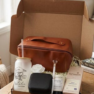 May include: A graduation gift box containing a brown leather wash bag, a white tumbler with a graduation cap design, a black jewellery box, a diamond-topped pen, a candle, a white rose, and a chocolate bar. The tumbler reads "Natalie Class of 2026."
