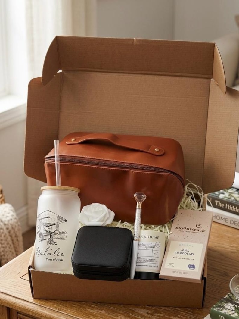 May include: A graduation gift box containing a brown leather wash bag, a white tumbler with a graduation cap design, a black jewellery box, a diamond-topped pen, a candle, a white rose, and a chocolate bar. The tumbler reads "Natalie Class of 2026."