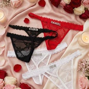 Peut inclure: Ensemble de strings rouges, noirs et blancs, avec bordures en dentelle et ornements en strass. Les noms "CHRIS", "DANIEL" et "BRIDE" sont affich&eacute;s. Dispos&eacute;s sur une surface satin&eacute;e rose avec des roses et des bougies.