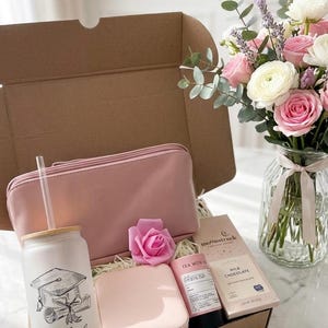 May include: A gift box with a pink theme, containing a tumbler with a graduation cap design, a pink cosmetic bag, a jewellery box, a rose, tea, and chocolate. A bouquet of pink and white flowers sits nearby.