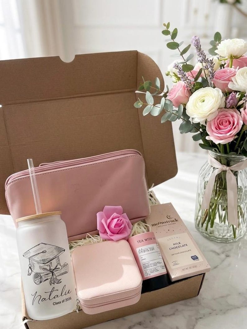 May include: A gift box with a pink theme, containing a tumbler with a graduation cap design, a pink cosmetic bag, a jewellery box, a rose, tea, and chocolate. A bouquet of pink and white flowers sits nearby.