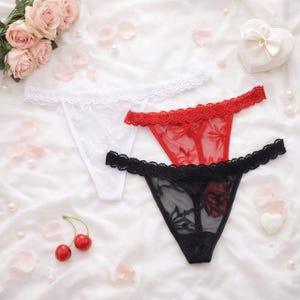 Peut inclure: Trois strings d&eacute;licats en blanc, rouge et noir, chacun avec une bordure en dentelle. Le tissu transparent r&eacute;v&egrave;le des motifs floraux. La lingerie est dispos&eacute;e sur une surface blanche textur&eacute;e avec des p&eacute;tales de rose et des perles.