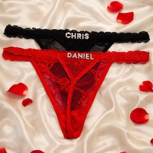 May include: Two thong underwear, one black and one red, with lace detailing. The black thong has the name "CHRIS" spelled out in rhinestones, and the red thong has "DANIEL". Red rose petals are scattered around the underwear.